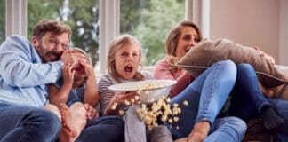 Frightened Family Sitting On Sofa At Home Watching Horror Movie On Tv With Popcorn