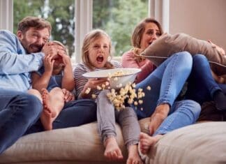 Frightened Family Sitting On Sofa At Home Watching Horror Movie On Tv With Popcorn