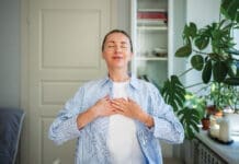 Relaxed Smiling Woman Meditates With Hands On Chest, Practice Breathing Exercises At Home. Self Care
